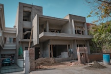 A two-story building is under construction, featuring a concrete structure with unfinished walls and open windows. Construction materials such as sand, bricks, and a stack of bags are visible in the front yard, along with some makeshift scaffolding. There is a blue barrel on the ground and some greenery on the right side of the image.
