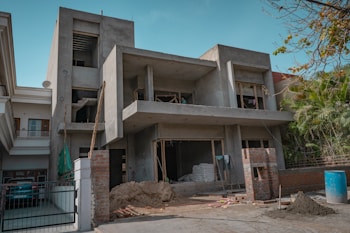 A two-story building is under construction, featuring a concrete structure with unfinished walls and open windows. Construction materials such as sand, bricks, and a stack of bags are visible in the front yard, along with some makeshift scaffolding. There is a blue barrel on the ground and some greenery on the right side of the image.