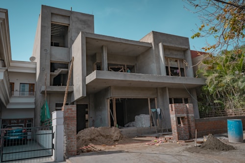 A two-story building is under construction, featuring a concrete structure with unfinished walls and open windows. Construction materials such as sand, bricks, and a stack of bags are visible in the front yard, along with some makeshift scaffolding. There is a blue barrel on the ground and some greenery on the right side of the image.