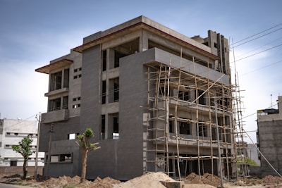 A multi-story building under construction is surrounded by wooden scaffolding. The concrete structure has several windows, and piles of dirt are visible around the building. The sky is clear with a few scattered clouds, and some greenery is present in the form of partially leafless trees.