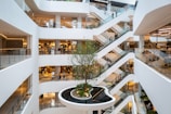Multiple levels of an indoor shopping mall with white walls and various stores on each floor. There are escalators crisscrossing between different levels, and a large tree is planted in the center atrium.