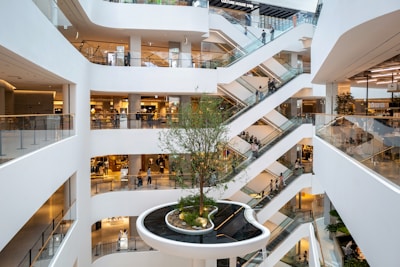 Multiple levels of an indoor shopping mall with white walls and various stores on each floor. There are escalators crisscrossing between different levels, and a large tree is planted in the center atrium.