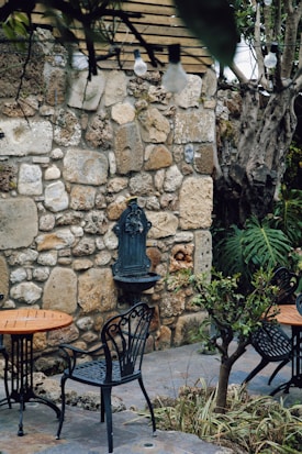 A rustic outdoor setting with wrought iron chairs and tables positioned on a stone-paved patio. The area is adorned with a weathered stone wall featuring a decorative water fountain. Overhanging light bulbs add an eclectic touch, while nearby plants and a small tree contribute to the garden ambiance.