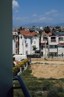 Residential neighborhood with white buildings featuring red roofs, surrounded by palm trees and some fenced gardens. The view includes a clear blue sky with a few clouds and distant mountains on the horizon.