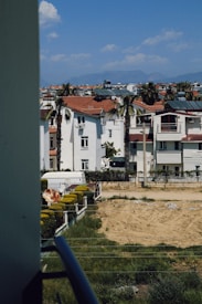 Residential neighborhood with white buildings featuring red roofs, surrounded by palm trees and some fenced gardens. The view includes a clear blue sky with a few clouds and distant mountains on the horizon.