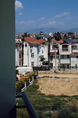 Residential neighborhood with white buildings featuring red roofs, surrounded by palm trees and some fenced gardens. The view includes a clear blue sky with a few clouds and distant mountains on the horizon.