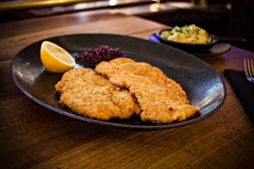 A black plate features two large breaded schnitzels accompanied by a lemon wedge and a small serving of cranberry sauce. In the background, a dish with a creamy potato salad garnished with herbs is slightly out of focus. The setting is on a wooden table, creating a rustic and warm dining atmosphere.