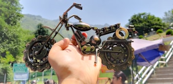 A hand is holding a small, intricately crafted model of a motorcycle made from various metal components, with a blurred outdoor background including greenery and a canopy.