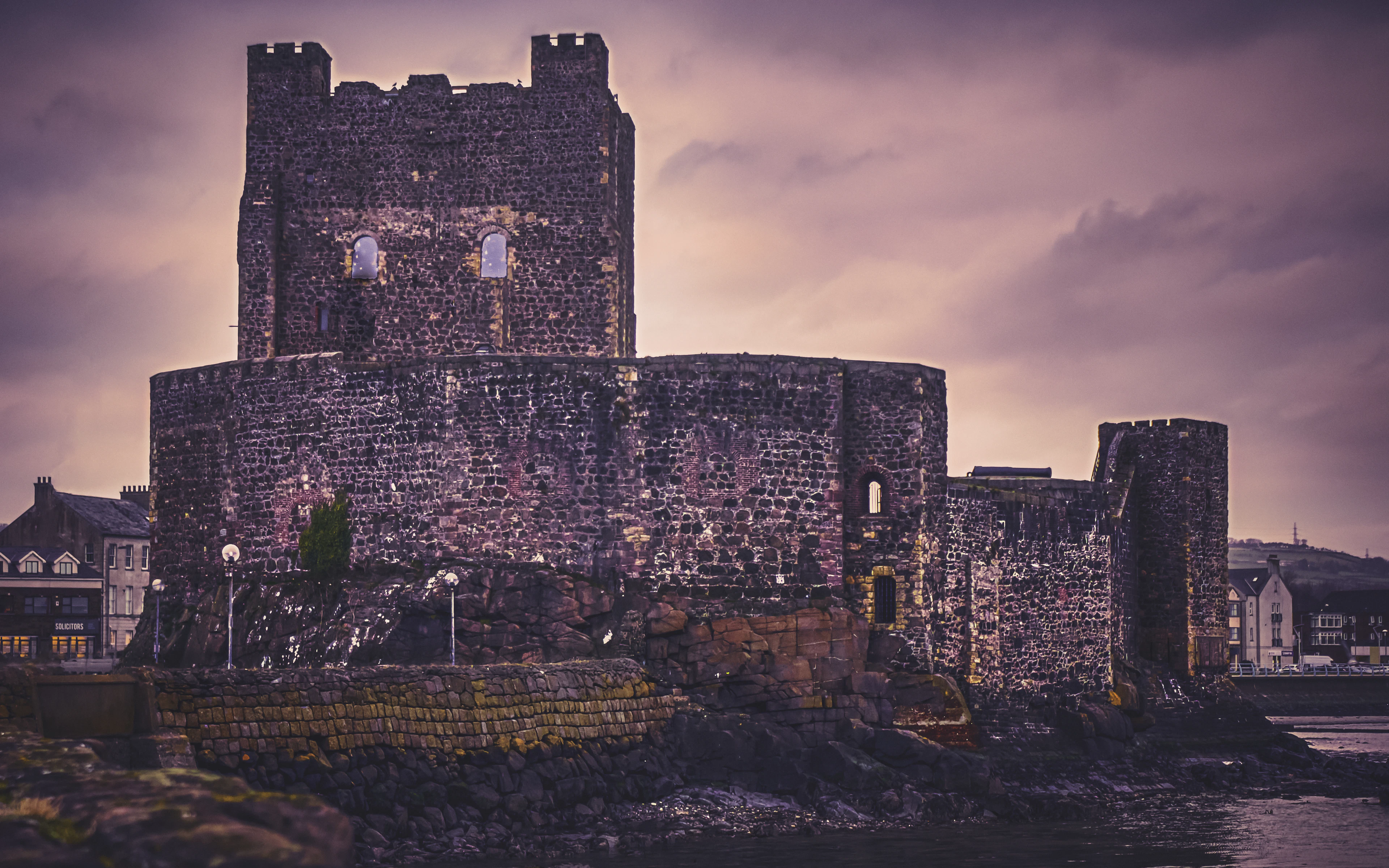 A medieval stone castle with round towers on a rocky coast under a dramatic dusk sky.