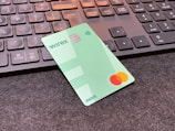 A green debit card with contactless payment symbol and a Mastercard logo is placed on a dark surface in front of a black computer keyboard. The keyboard keys have a soft orange backlight.