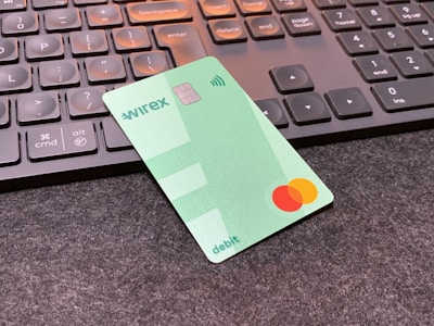 A green debit card with contactless payment symbol and a Mastercard logo is placed on a dark surface in front of a black computer keyboard. The keyboard keys have a soft orange backlight.