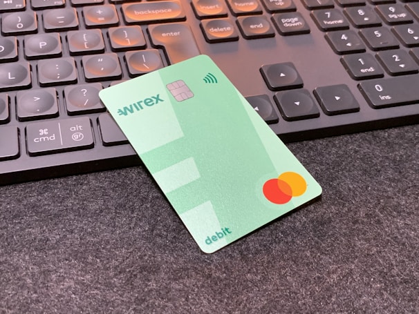 A green debit card with contactless payment symbol and a Mastercard logo is placed on a dark surface in front of a black computer keyboard. The keyboard keys have a soft orange backlight.