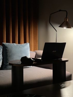A cozy indoor setting with dim lighting, featuring a laptop on a small table in front of a sofa. Beside the laptop, there is a pair of headphones. A modern floor lamp provides soft light, highlighting the laptop and part of the sofa. Behind the sofa are brown curtains and a blue cushion.