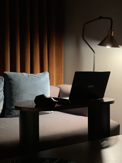 A cozy indoor setting with dim lighting, featuring a laptop on a small table in front of a sofa. Beside the laptop, there is a pair of headphones. A modern floor lamp provides soft light, highlighting the laptop and part of the sofa. Behind the sofa are brown curtains and a blue cushion.