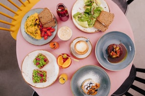 A round table with a pink tablecloth is set with a variety of colorful dishes, including an omelet with cherry tomatoes and bread, an avocado salad with leafy greens, a stack of pancakes garnished with berries, and a dessert covered in a glossy sauce. Drinks include a latte, fresh juice in small glasses, and a vibrant cocktail with an orange slice. The setting includes yellow and black chairs, contributing to an inviting dining atmosphere.