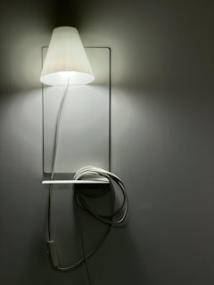 A simple, wall-mounted lamp emits a soft glow in a dimly lit room. Attached is a white cord loosely wrapped around a small horizontal ledge beneath the lamp.