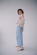 A young person is posing in a simplistic setting with a white background. They are wearing a light pink off-the-shoulder top, a beige jacket draped over their arms, light blue high-waisted jeans, and white sneakers with orange accents. The individual has a short, curly hairstyle with streaks of purple or red hair dye. Their makeup is bold with bright pink eyeshadow.
