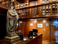 A richly decorated room with wooden paneling and a two-level structure featuring bookshelves on the upper level. A large statue of a robed figure holding a child stands prominently in the foreground. Below, there is a desk with a computer and a tufted leather chair, complemented by framed artwork on the walls.