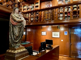 A richly decorated room with wooden paneling and a two-level structure featuring bookshelves on the upper level. A large statue of a robed figure holding a child stands prominently in the foreground. Below, there is a desk with a computer and a tufted leather chair, complemented by framed artwork on the walls.
