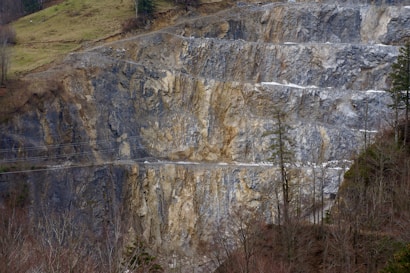 A steep rock face of a quarry with exposed layers of gray and beige stone. Sparse vegetation is visible at the top and bottom of the slope, with a few tall, thin trees on the right side.