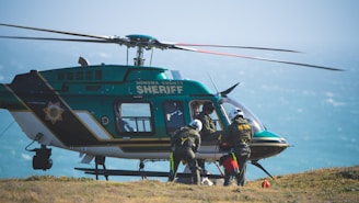 A sheriff's helicopter is stationed on the ground with its rotors still. Several individuals dressed in rescue gear are preparing to board or have just exited the helicopter. The background shows a hazy sky over a body of water.