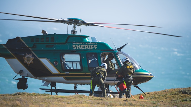 A sheriff's helicopter is stationed on the ground with its rotors still. Several individuals dressed in rescue gear are preparing to board or have just exited the helicopter. The background shows a hazy sky over a body of water.
