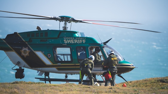 A sheriff's helicopter is stationed on the ground with its rotors still. Several individuals dressed in rescue gear are preparing to board or have just exited the helicopter. The background shows a hazy sky over a body of water.