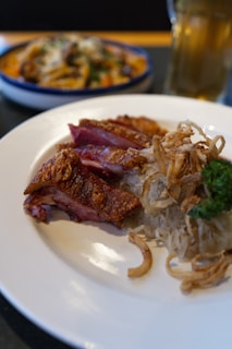 A white plate holds crispy sliced duck, garnished with cooked onions and a sprig of parsley. Nearby, there is another dish in the background, slightly out of focus, along with a tall glass of beer.