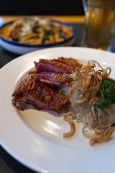 A white plate holds crispy sliced duck, garnished with cooked onions and a sprig of parsley. Nearby, there is another dish in the background, slightly out of focus, along with a tall glass of beer.