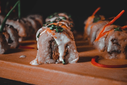 Several sushi rolls are neatly arranged on a wooden board, topped with creamy white sauce and garnished with sliced green onions and orange strips. The rolls have a rich texture and appear to be stuffed with a deep-fried filling.