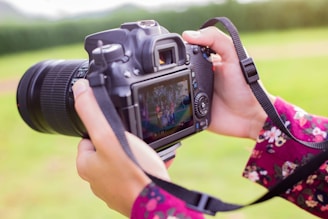 Hands are holding a digital camera with a large lens. The camera is displaying a photo on its screen, featuring a group of people gathered together. The person holding the camera is wearing a floral-patterned garment. The background is a blurry green landscape, indicating an outdoor setting.