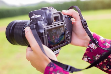 Hands are holding a digital camera with a large lens. The camera is displaying a photo on its screen, featuring a group of people gathered together. The person holding the camera is wearing a floral-patterned garment. The background is a blurry green landscape, indicating an outdoor setting.