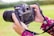 Hands are holding a digital camera with a large lens. The camera is displaying a photo on its screen, featuring a group of people gathered together. The person holding the camera is wearing a floral-patterned garment. The background is a blurry green landscape, indicating an outdoor setting.