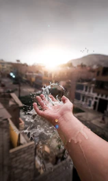 A hand is extended into the air, capturing water droplets that splash upwards against a blurred urban background. The sun is setting or rising on the horizon, casting a warm glow over the scene. Birds are flying in the distant sky.