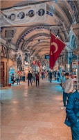 An indoor market with arched ceilings adorned with decorative patterns and hanging Turkish flags. Numerous people are walking and browsing the shops that line both sides of the walkway. The floor is made of large tiles, and shop signs are visible in various places, contributing to a bustling, vibrant atmosphere.