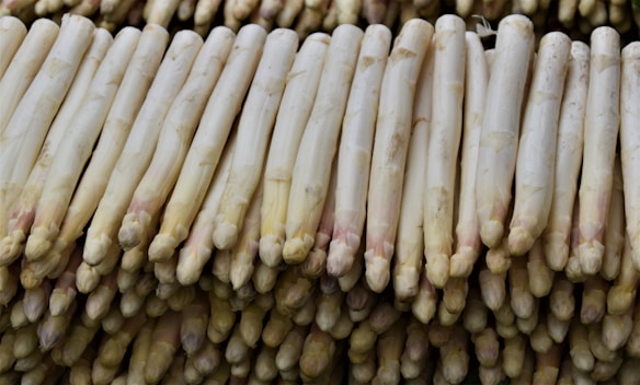 A large pile of white asparagus stacked neatly. The spears are arranged horizontally, showing their smooth, light-colored surface with slight hints of pink or purple at the tips.