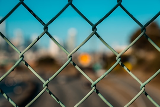 A chain-link fence in the foreground with an out-of-focus urban landscape in the background. The sky is clear and blue.
