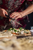 A person wearing a floral shirt is preparing a homemade pizza, using a knife to sprinkle fresh herbs onto it. The pizza is topped with ingredients such as basil leaves, cheese, and zucchini slices. The background is slightly out of focus, giving a warm and intimate kitchen setting.