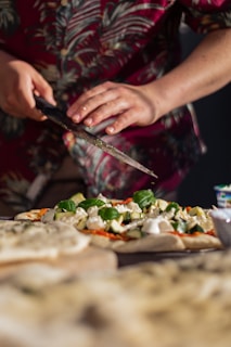 A person wearing a floral shirt is preparing a homemade pizza, using a knife to sprinkle fresh herbs onto it. The pizza is topped with ingredients such as basil leaves, cheese, and zucchini slices. The background is slightly out of focus, giving a warm and intimate kitchen setting.