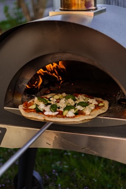A pizza is being baked in a wood-fired outdoor oven. The pizza features toppings like fresh basil leaves, cheese, and zucchini slices on a tomato base. The fire inside the oven is visible, providing a warm, cozy ambiance. A metal pizza peel is being used to place the pizza into the oven.