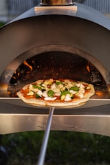 A pizza topped with fresh vegetables and cheese is placed on a wooden pizza peel, being inserted into a wood-fired oven. The flames inside the oven create a warm, inviting atmosphere with a glow reflecting on the pizza dough.