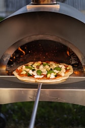 A pizza topped with fresh vegetables and cheese is placed on a wooden pizza peel, being inserted into a wood-fired oven. The flames inside the oven create a warm, inviting atmosphere with a glow reflecting on the pizza dough.