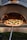 A pizza topped with fresh vegetables and cheese is placed on a wooden pizza peel, being inserted into a wood-fired oven. The flames inside the oven create a warm, inviting atmosphere with a glow reflecting on the pizza dough.