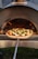 A pizza topped with fresh vegetables and cheese is placed on a wooden pizza peel, being inserted into a wood-fired oven. The flames inside the oven create a warm, inviting atmosphere with a glow reflecting on the pizza dough.