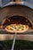 A pizza topped with fresh vegetables and cheese is placed on a wooden pizza peel, being inserted into a wood-fired oven. The flames inside the oven create a warm, inviting atmosphere with a glow reflecting on the pizza dough.