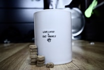 A white mug is placed on a wooden surface. The mug features the text 'Live, Love + Eat Treats' with a small paw print below the text. Beside the mug, there are stacks of round, beige biscuits arranged in piles. The background is out of focus, featuring dimly lit kitchen utensils or appliances.
