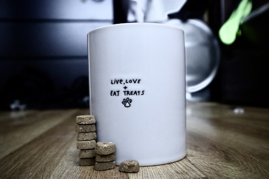 A white mug is placed on a wooden surface. The mug features the text 'Live, Love + Eat Treats' with a small paw print below the text. Beside the mug, there are stacks of round, beige biscuits arranged in piles. The background is out of focus, featuring dimly lit kitchen utensils or appliances.