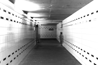 A tiled underground passageway with fluorescent lighting. The walls are lined with white tiles featuring a black square pattern near the top and bottom. An SOS phone is mounted on the left wall, and there are handrails on the right. The floor is made of rectangular bricks, and the passage leads to an exit at the far end.