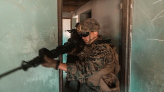 A soldier in full combat gear, including helmet and goggles, is holding a rifle and moving tactically through a narrow, dimly-lit passageway. The walls are painted a worn teal color, and the light in the space is subdued, adding to the tense atmosphere.