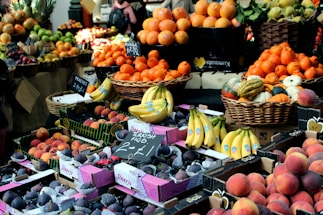 A vibrant market scene with a wide variety of fruits on display. Baskets and boxes are filled with oranges, bananas, figs, various types of squash, and peaches. Signs indicate prices and labels for the produce, creating a lively and colorful assortment.